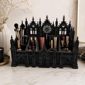 Peut inclure: Un organisateur de maquillage noir de style gothique, en forme de cathédrale, avec des détails architecturaux complexes. Il contient plusieurs compartiments pour ranger des articles de maquillage, tels que des pinceaux et des rouges à lèvres. Le fond est une surface en marbre.