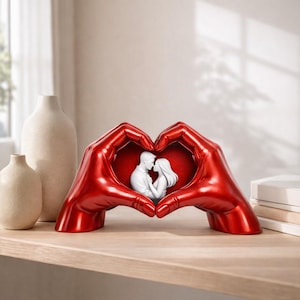 May include: A red sculpture of two hands forming a heart shape, framing a white statue of a couple kissing. The sculpture is on a wooden shelf, with two beige vases in the background. The hands have a glossy finish.