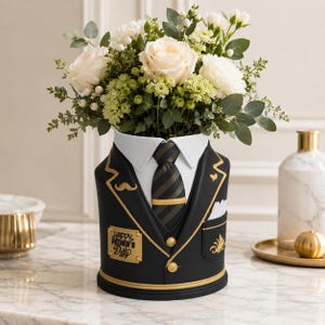 May include: A black vase shaped like a suit jacket, with a white collar and striped tie, holding a bouquet of white and green flowers. The vase has gold accents and the text "Happy Father's Day".