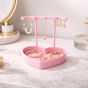May include: A pink heart-shaped jewelry organizer with a stand for necklaces and earrings. The organizer holds bracelets and rings. The jewelry is gold and pearl. The background is a white marble surface.