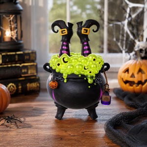 Puede incluir: Un caldero negro con líquido verde burbujeante y piernas de bruja, una decoración de Halloween. El caldero tiene un pequeño amuleto de calabaza y una botella de poción morada. Una calabaza tallada y una telaraña están en el fondo.