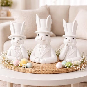 May include: Three white ceramic Easter bunny figurines wearing top hats and tuxedos, arranged on a woven mat with pastel-colored Easter eggs and baby's breath. The bunnies are on a white table in front of a beige sofa.