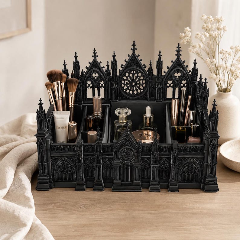 Puede incluir: Un organizador de maquillaje negro de estilo g&oacute;tico con m&uacute;ltiples compartimentos. El organizador tiene forma de catedral, con intrincados detalles arquitect&oacute;nicos. Contiene brochas de maquillaje, tubos, frascos y otros productos de belleza.