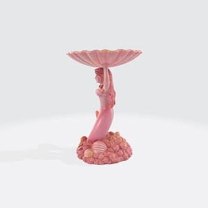 Pu&ograve; includere: Una statuetta di sirena rosa che tiene una grande conchiglia smerlata. La sirena ha lunghi capelli e una coda squamata. La base &egrave; un gruppo di coralli e conchiglie. Un elemento decorativo.