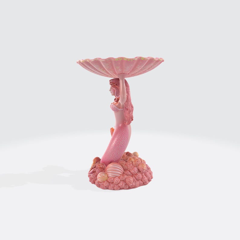 Pu&ograve; includere: Una statuetta di sirena rosa che tiene una grande conchiglia smerlata. La sirena ha lunghi capelli e una coda squamata. La base &egrave; un gruppo di coralli e conchiglie. Un elemento decorativo.