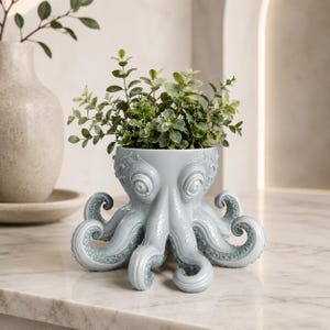 Può includere: Un vaso a forma di polpo blu chiaro con tentacoli e occhi dettagliati, contenente piante verdi. Il vaso in ceramica è posto su una superficie di marmo, offrendo un design decorativo unico per la casa.