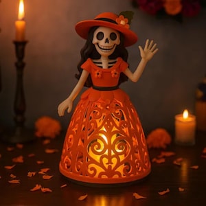 Puede incluir: Figura naranja del Día de Muertos de una mujer con rostro de esqueleto, sombrero y vestido. El vestido tiene un diseño calado decorativo que brilla con luz interna. La figura es un artículo decorativo.