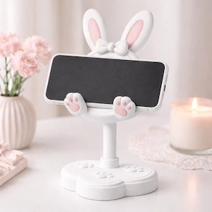 May include: A white bunny-themed phone stand with pink accents. The stand features bunny ears, a bow, and paw-shaped details. It holds a black smartphone, perfect for hands-free viewing. The base has paw prints.