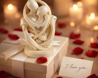 Puede incluir: Una escultura crema de dos figuras abrazadas con un recorte en forma de corazón. La escultura se encuentra sobre una caja de regalo atada con una cinta crema. Pétalos de rosa rojos y una tarjeta con "I love you" están esparcidos.