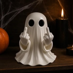 Könnte beinhalten: Weiße Keramik-Geisterfigur mit schwarzen Augenlöchern, die mit beiden Händen den Mittelfinger zeigen. Die Figur steht auf einer Holzoberfläche, mit Kürbis und Kerzen im Hintergrund. Halloween-Dekoration.