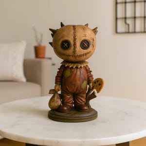 Puede incluir: Figura decorativa de un personaje con cabeza cosida, ojos de botón y pelo puntiagudo. La figura viste un atuendo marrón con parches y sostiene un saco y una piruleta. La figura está sobre una base redonda.