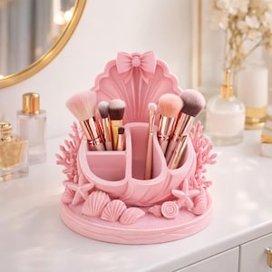 May include: A pink seashell-shaped makeup brush holder, adorned with a bow, coral, seashells, and starfish. The holder contains makeup brushes with gold handles. This decorative item is designed to organize and display makeup brushes in a stylish manner.