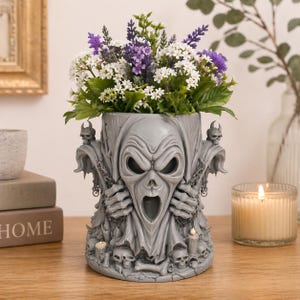 May include: A grey, decorative planter with a screaming skull design, filled with white and purple flowers. The planter has a detailed, gothic aesthetic, with skulls and chains. A lit candle sits nearby.