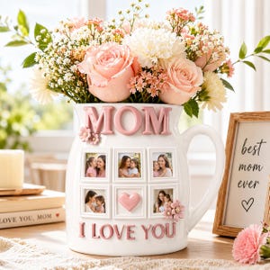 Può includere: Vaso in ceramica bianca con la scritta "MOM" in lettere rosa, che contiene un bouquet di fiori rosa e bianchi. Il vaso ha cornici per foto e le parole "I LOVE YOU" in rosa. Un cartello incorniciato recita "best mom ever".
