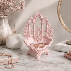 May include: A light pink, ornate jewelry dish with a gothic arch design. The dish holds gold jewelry, including a ring and necklace. The decorative piece is on a marble surface.
