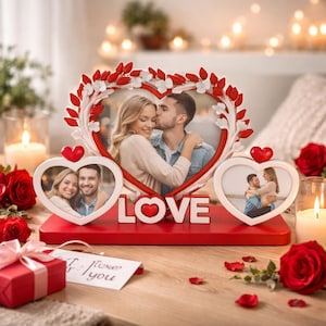 以下が含まれることがあります： 白い花の模様と「LOVE」の文字が入った赤いハート型のフォトフレーム。フレームにはカップルの写真が3枚入っています。赤いギフトボックスとバラがテーブルに置かれています。