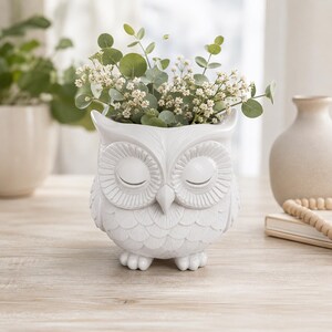 Op de afbeelding: Een witte keramische uilvormige plantenbak met gesloten ogen en gedetailleerde vedertexturen. De uil is gevuld met groene eucalyptusbladeren en kleine witte bloemen, wat een decoratieve houder vormt voor kamerplanten of bloemstukken.