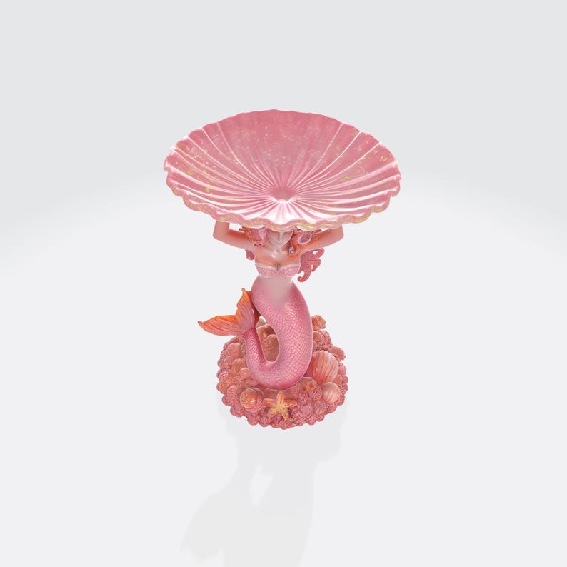 Pu&ograve; includere: Una statuina di sirena rosa che tiene una grande conchiglia smerlata. La sirena ha i capelli rosa e una coda di pesce, ed &egrave; su una base con conchiglie e stelle marine. &Egrave; un oggetto decorativo.
