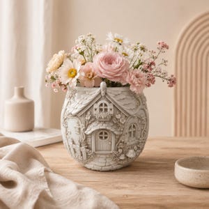 May include: A light gray ceramic vase shaped like a small cottage with detailed relief carvings of a door, windows, and a picket fence, filled with a colorful arrangement of white, pink, and cream-colored flowers including daisies and roses.