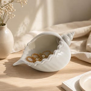 Könnte beinhalten: Eine weiße, muschelförmige Schmuckschale aus Keramik. Die Schale enthält goldene Ringe und Halsketten mit Perlen. Die Muschel hat eine strukturierte Oberfläche und einen gewellten Rand. Im Hintergrund befindet sich eine kleine Vase mit Blumen.