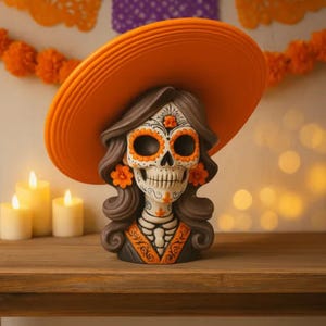 Può includere: Una statuetta del Giorno dei Morti con un teschio con intricati disegni floreali e geometrici. Il teschio indossa un cappello arancione e ha accenti floreali arancioni. La statuetta è su una superficie di legno con candele accese e carta arancione decorativa.