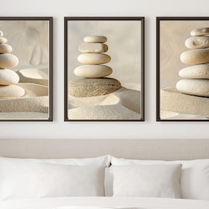 Puede incluir: Tres impresiones enmarcadas que muestran pilas de piedras lisas y claras sobre arena. Las piedras están dispuestas en formaciones equilibradas, creando una sensación de tranquilidad. La obra de arte está sobre un fondo neutro, adecuada para la decoración del hogar.