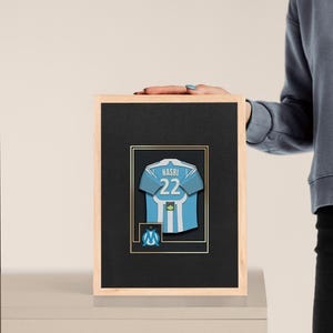 May include: Framed artwork featuring a light blue and white soccer jersey with the name "NASRI" and the number "22". The jersey is set against a black background with a gold border, inside a wooden frame. The team logo is visible.