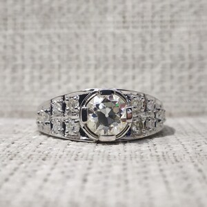 May include: A silver-toned ring featuring a large, round, faceted center stone held by a bezel setting. The band is accented with two rows of three smaller, round, faceted stones on each side, creating a symmetrical design with a polished finish.