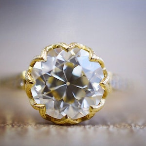 Puede incluir: Primer plano de un anillo de oro con una gema grande, clara y facetada. El anillo tiene un engaste de oro ornamentado con un diseño festoneado alrededor de la gema. La banda del anillo también es de oro y tiene pequeños detalles.