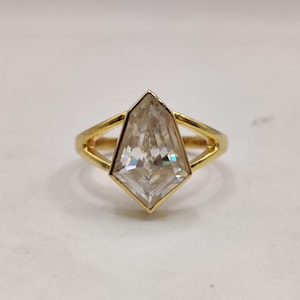 May include: A gold ring featuring a large, clear, kite-shaped faceted gemstone set in a bezel setting. The band of the ring splits into two thin, polished gold arms that connect to the sides of the central stone, creating a geometric design.