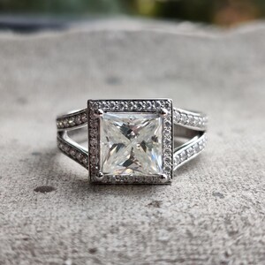 May include: A silver ring with a large, square, clear gemstone at the center, surrounded by smaller, clear stones. The band splits into two sections on each side, also adorned with small, clear stones. The ring is set against a neutral background.