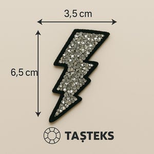 Könnte beinhalten: Ein schwarzer und silberner Blitz-Aufnäher, ca. 3,5 cm breit und 6,5 cm hoch. Der Aufnäher ist mit silbernen Strasssteinen verziert und hat einen schwarzen Rand. Der Text "TAŞTEKS" ist darunter aufgedruckt.