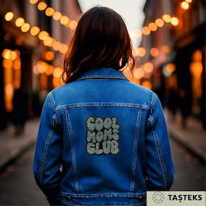 May include: A blue denim jacket with silver glitter text reading "COOL MOMS CLUB" on the back. The jacket features a classic design with button fastenings and pockets. The image is taken from behind, showcasing the jacket's details.