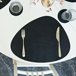 May include: A black leather placemat with a silver fork and knife set on a white table. A clear glass and a small bowl are also on the table. The table is set for a meal.