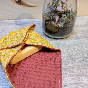 Puede incluir: Un jabonera con estampado geom&eacute;trico amarillo y blanco con interior color &oacute;xido, que contiene una pastilla de jab&oacute;n. Un terrario de cristal con una peque&ntilde;a planta est&aacute; en el fondo. El soporte se cierra con un broche amarillo.
