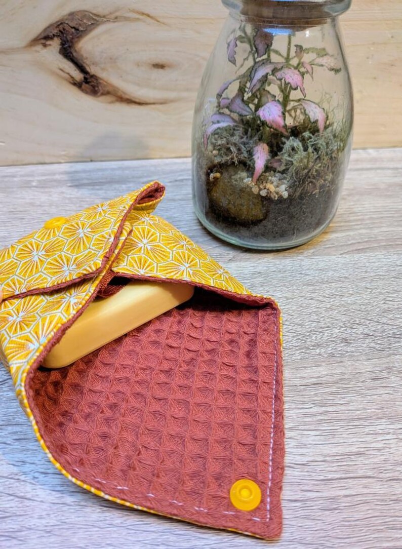 Puede incluir: Un jabonera con estampado geom&eacute;trico amarillo y blanco con interior color &oacute;xido, que contiene una pastilla de jab&oacute;n. Un terrario de cristal con una peque&ntilde;a planta est&aacute; en el fondo. El soporte se cierra con un broche amarillo.