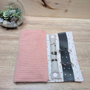 Puede incluir: Un estuche enrollable de joyería de pana rosa con interior estampado floral. El rollo tiene secciones para anillos y pendientes. El rollo está abierto, mostrando anillos y pendientes. El rollo está sobre una superficie de madera.