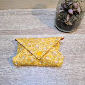 Puede incluir: Una cartera de tela amarilla con un patr&oacute;n geom&eacute;trico de panal. La cartera est&aacute; doblada y asegurada con un bot&oacute;n amarillo. El ribete interior es rojo. Un peque&ntilde;o terrario con plantas rosas est&aacute; en el fondo.