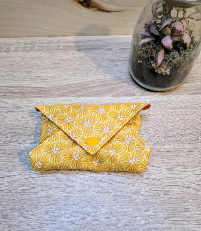 Puede incluir: Una cartera de tela amarilla con un patr&oacute;n geom&eacute;trico de panal. La cartera est&aacute; doblada y asegurada con un bot&oacute;n amarillo. El ribete interior es rojo. Un peque&ntilde;o terrario con plantas rosas est&aacute; en el fondo.