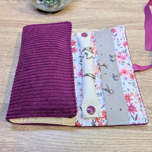 Puede incluir: Un estuche para joyas de pana morada con un forro interior floral de flores rosas y rojas. El estuche incluye un soporte para anillos de color crema con cierre a presión y una tira de cuero gris con agujeros para pendientes, sujeto por una cinta morada.