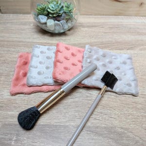 Op de afbeelding: Een make-up kwast en een wenkbrauwkam met grijze handvatten en gouden accenten worden getoond met drie zachte, getextureerde gezichtsdoekjes in roze en wit. Een glazen terrarium met vetplanten en stenen staat op de achtergrond.