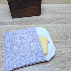 Puede incluir: Una bolsa de tela cuadrada con un patrón geométrico de estrellas en color violeta claro y forro interior blanco. La bolsa se asegura con un pequeño botón a presión violeta y contiene una pastilla de jabón amarilla rectangular en la solapa.