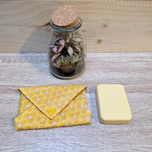 May include: A glass terrarium with a cork lid, containing a small plant with pink and green leaves. A yellow fabric pouch with a geometric pattern and a yellow button closure sits next to a rectangular bar of soap.