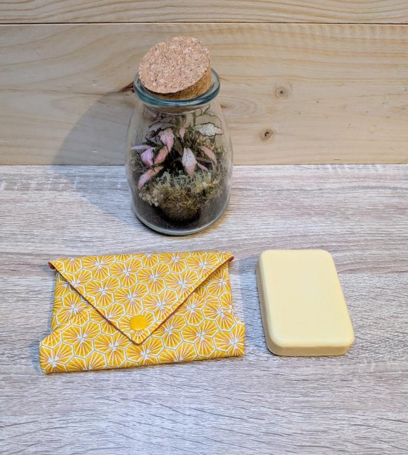 Puede incluir: Un terrario de vidrio con tapa de corcho, que contiene una peque&ntilde;a planta con hojas rosas y verdes. Una funda de tela amarilla con un patr&oacute;n geom&eacute;trico y cierre de bot&oacute;n amarillo se encuentra junto a una pastilla de jab&oacute;n rectangular.
