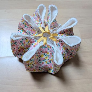 Könnte beinhalten: Ein Stoffbeutel mit Kordelzug und einem Blumenmuster aus rosa, gelben und grünen Blüten auf weißem Grund. Das Innere ist mit weißem Frottee gefüttert und die Öffnung wird durch gelbe Gummibänder und eine weiße Kordel zusammengehalten.