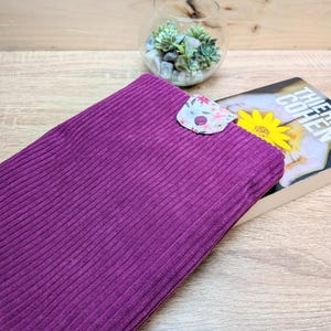 Puede incluir: Una funda de pana de color morado intenso con tejido de textura acanalada. La funda tiene una pequeña solapa de cierre redondeada en la parte superior, decorada con un estampado floral claro y un botón a presión morado centrado.