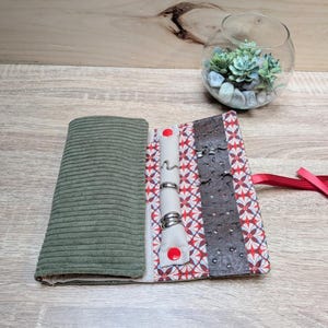 Puede incluir: Un estuche para joyas de tela con exterior de pana verde oliva e interior con estampado geométrico multicolor. Incluye un soporte para anillos de color crema con botones a presión rojos y una tira texturizada de color marrón oscuro.