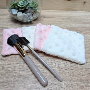 Op de afbeelding: Een make-up kwast met een gouden ferrule en een taupekleurige handgreep, een wenkbrauwborstel en verschillende kleine, getextureerde, roze en witte gezichtsdoekjes zijn gerangschikt op een houten oppervlak. Een glazen terrarium met vetplanten en stenen staat op de achtergrond.