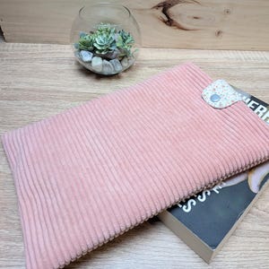 Puede incluir: Una funda de libro de pana rosa con un cierre de lengüeta con estampado floral. La funda está colocada sobre un libro, cuyo título es parcialmente visible. Un terrario de cristal con suculentas está en el fondo.