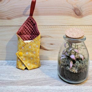 Puede incluir: Una bolsa de tela amarilla con un patr&oacute;n de panal y un cierre de bot&oacute;n amarillo, junto a un terrario de vidrio con tapa de corcho. La bolsa tiene un interior burdeos y un lazo burdeos.